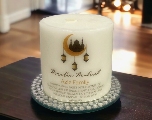 A white 8 cm candle with embellishments, with text saying Ramadan Mubarak followed by Aziz Family and a quote from the Quran. It is placed on a crystal base.